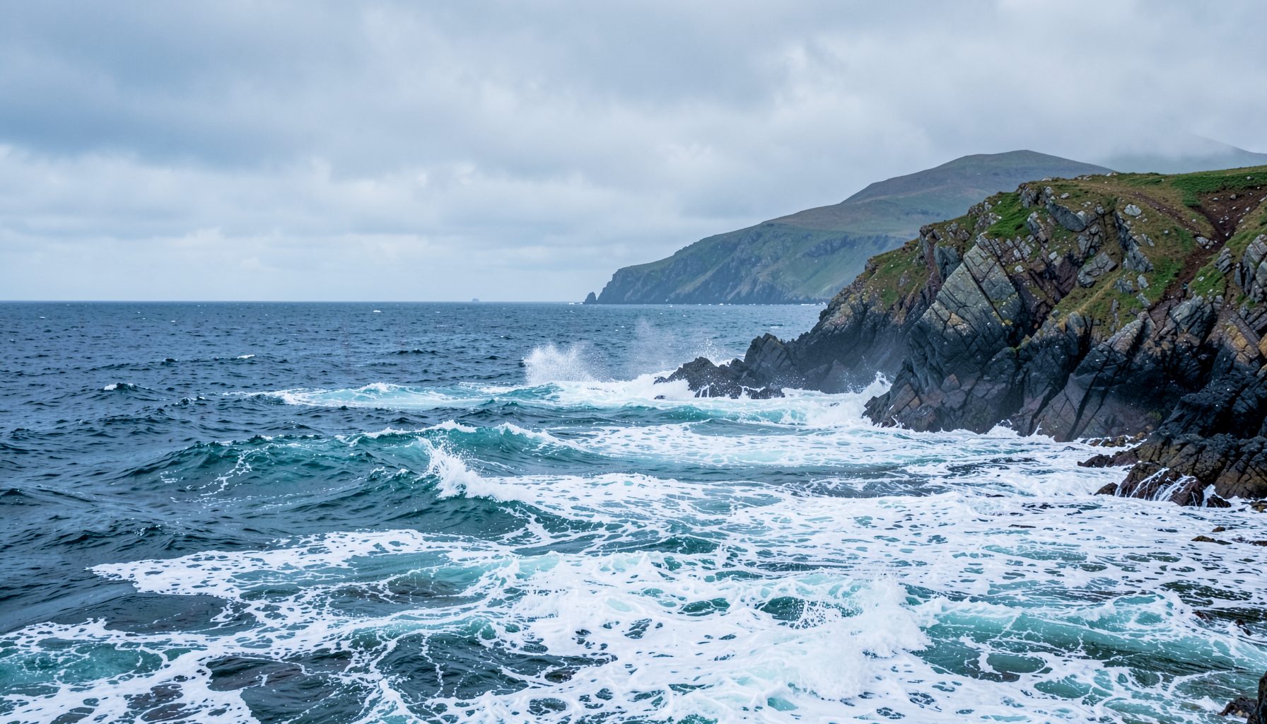 Côte rocheuse balayée par les vagues sous un ciel nuageux, tons gris-bleu et écume blanche